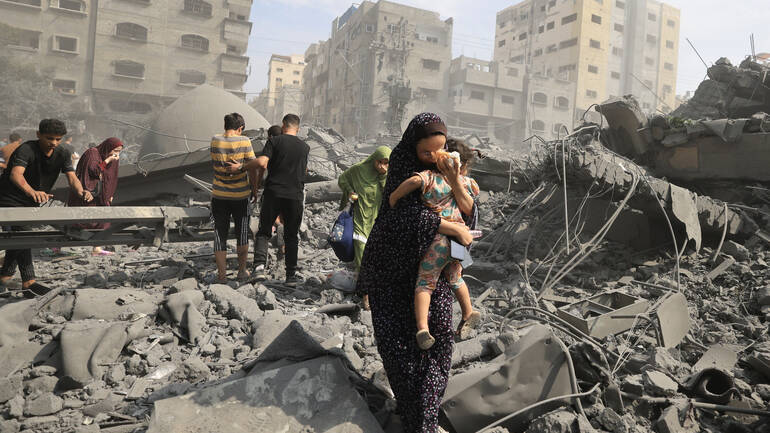 Eine Frau trägt ihr Kind durch eine von Angriffen zerstörte Straße in Gaza Stadt, im Hintergrund weitere Menschen.