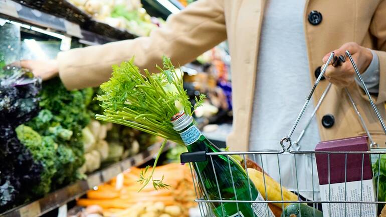 Eine Person steht mit Einkaufskorb vor dem Gemüseregal in einem Supermarkt.