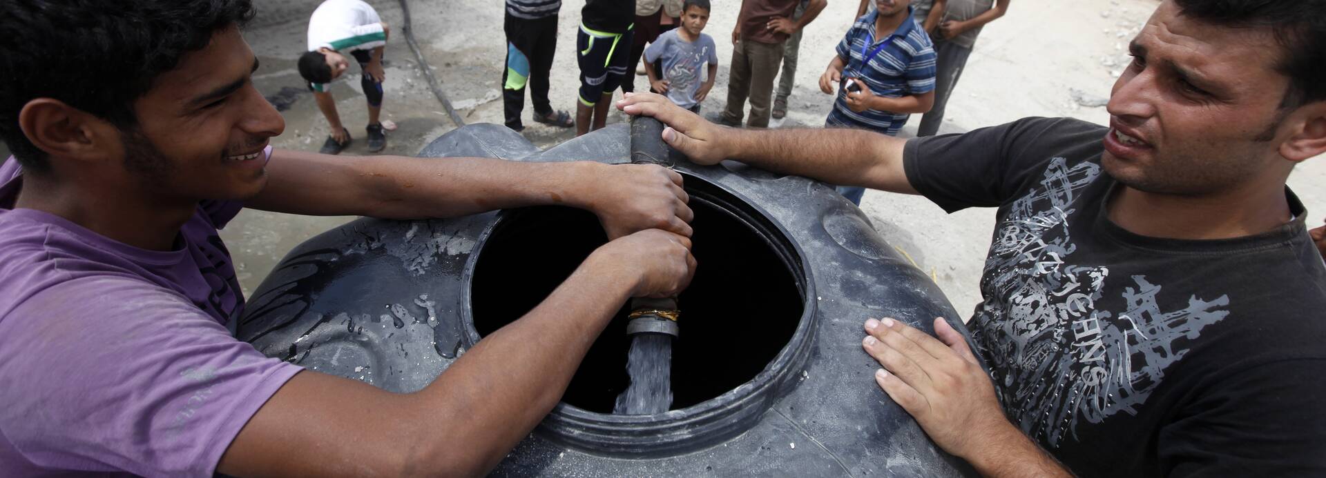 Zwei Männer füllen einen Wassertank mit frischem Wasser, im Hintergrund stehen weitere Menschen