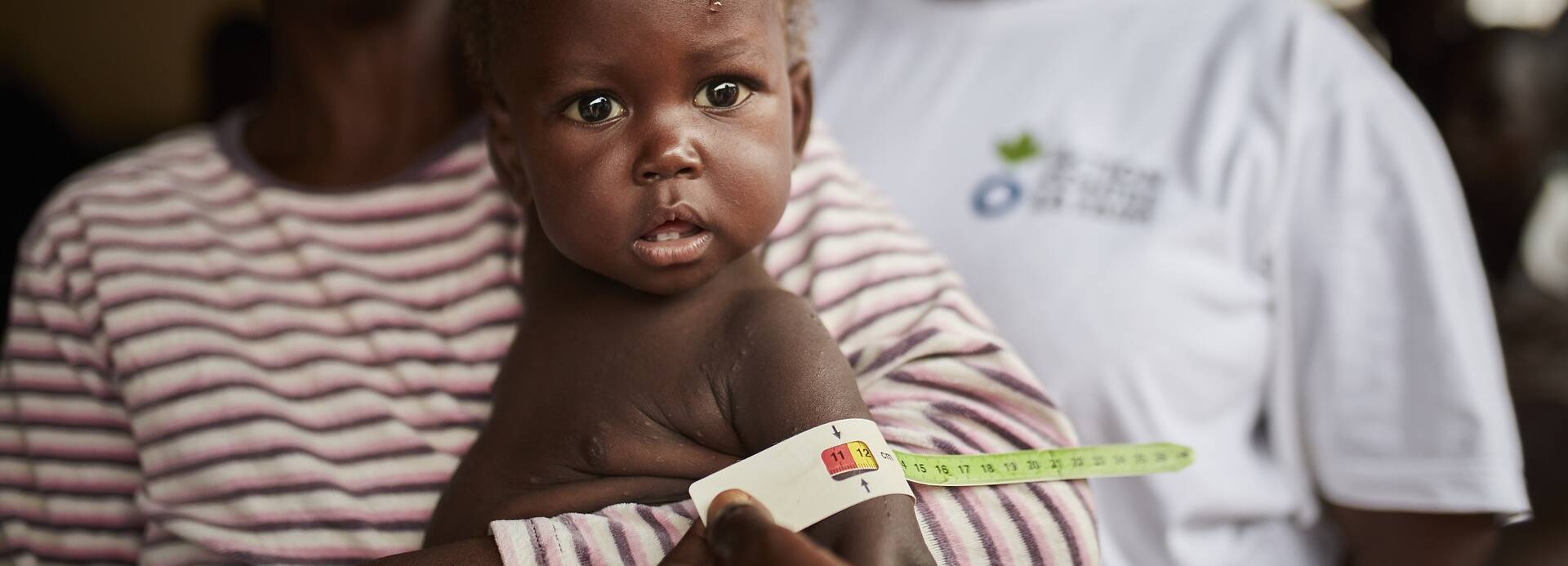 Ein Kind mit wird mit MUAC-Band auf Mangelernährung untersucht, während die Mutter es im Arm hält, im Hintergrund eine Mitarbeiterin von Aktion gegen den Hunger.