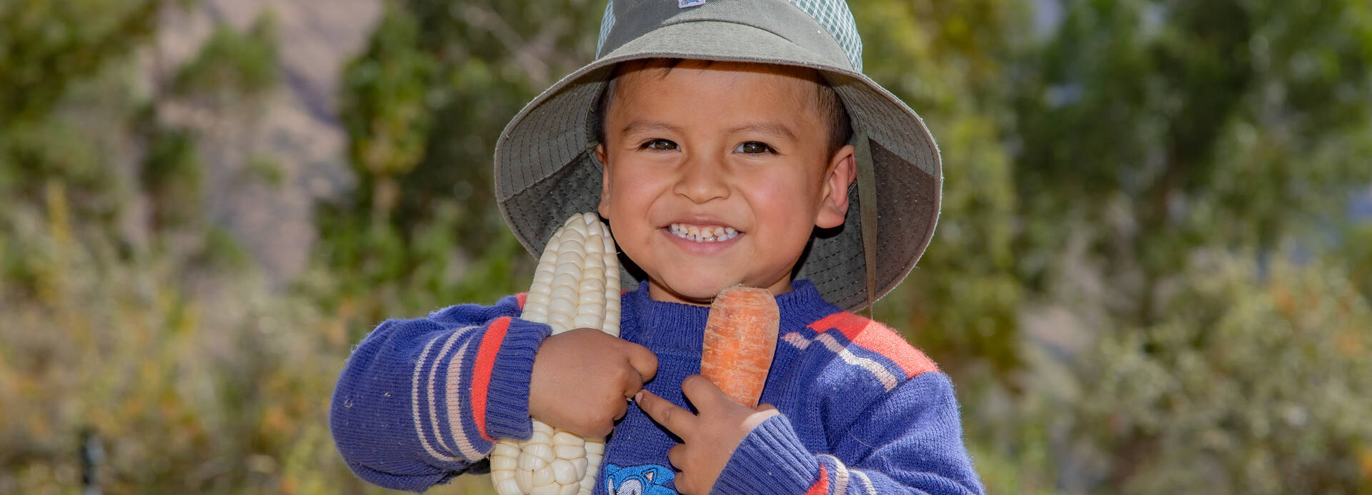 Ein Junge aus Peru hält einen Maiskolben und eine Karotte in seinen Händen und freut sich über die Ernte.