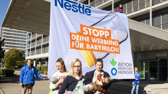 Mütter mit Babys stillen vor der Nestlé-Zentrale in Frankfurt am Main, im Hintergrund halten Aktivist*innen von Aktion gegen den Hunger ein Banner: Nestlé: Stopp deine Werbung für Babymilch!