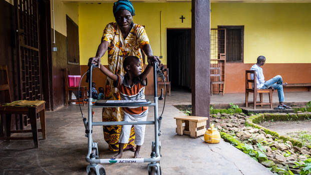 Dieubéni kann mithilfe eines Rollators endlich das Laufen lernen. Seine Großmutter stützt ihn dabei und hält seine Hände am Gehwagen fest, während sie durch den Innenhof des Therapiezentrums laufen. 