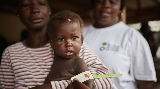 Ein Kind mit wird mit MUAC-Band auf Mangelernährung untersucht. Ein Kind mit wird mit MUAC-Band auf Mangelernährung untersucht, während die Mutter es im Arm hält, im Hintergrund eine Mitarbeiterin von Aktion gegen den Hunger.