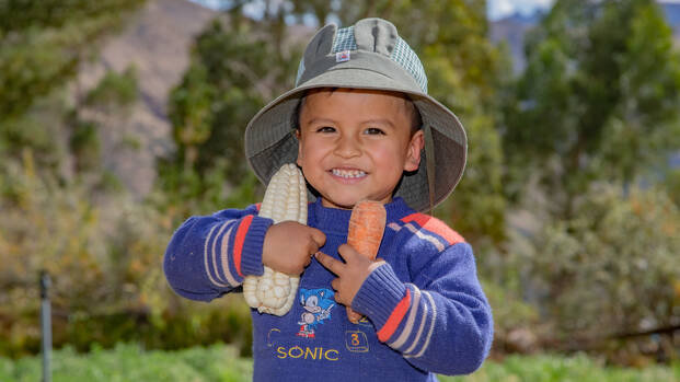 Ein Junge aus Peru hält einen Maiskolben und eine Karotte in seinen Händen und freut sich über die Ernte.